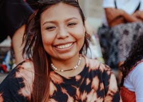 A smiling woman with long hair and eyeglasses sits outdoors, wearing a tie-dye shirt. Urban setting.